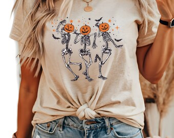 May include: Beige t-shirt with a Halloween design. The graphic depicts three dancing skeletons with pumpkin heads, surrounded by stars and bats. The shirt is knotted at the waist and worn with blue denim shorts.