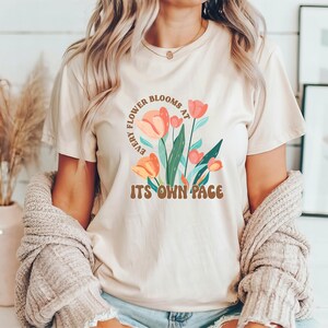 Könnte beinhalten: Beiges T-Shirt mit floralem Aufdruck. Das Design zeigt orange und rosa Tulpen mit grünen Blättern und den Text "EVERY FLOWER BLOOMS AT ITS OWN PACE" in braunen Buchstaben. Das Shirt wird mit einer beigen Strickjacke getragen.
