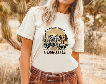 May include: A white t-shirt with a brown graphic of a woman riding a horse in a desert landscape. The text "Shoulda Been a Cowgirl" is printed on the shirt.