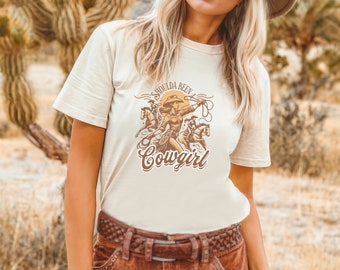 May include: A white t-shirt with a graphic of a woman riding a horse and the text "Shoulda Been a Cowgirl".