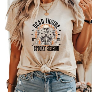 May include: Beige t-shirt with the text "DEAD INSIDE but it's SPOOKY SEASON" and a skeleton graphic. The skeleton is giving the peace sign. The shirt is knotted at the waist and paired with blue denim shorts.