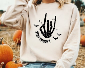 Może przedstawiać: Beżowa bluza z czarną grafiką szkieletowej dłoni w geście rock and roll. Pod spodem napis "STAY SPOOKY", z nietoperzami i gwiazdami. Odzież o tematyce jesiennej.