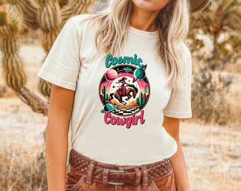 May include: A white t-shirt with a graphic design of a cowgirl riding a horse in front of a desert landscape with planets and stars. The text "Cosmic Cowgirl" is printed below the graphic.