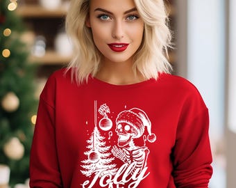 May include: A red sweatshirt featuring a white graphic of a skeleton in a Santa hat, holding a Christmas tree and baubles. The text "Jolly AF" is below. The sweatshirt is worn by a person with blonde hair and red lipstick.