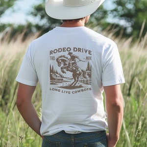 Puede incluir: Camiseta blanca con un gráfico marrón de un vaquero montando un caballo. El gráfico incluye el texto "Rodeo Drive", "TRD", "MRK" y "Long Live Cowboys".