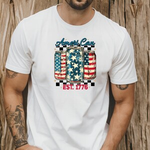 Puede incluir: Camiseta blanca con un gráfico de estilo vintage de tres latas con temática de la bandera estadounidense en rojo, blanco y azul. El texto "Ameri Can Est. 1776" está impreso debajo de las latas.