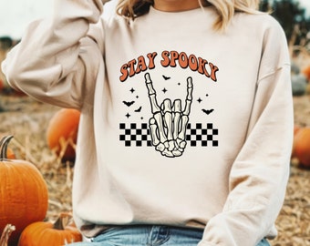 Może przedstawiać: Beżowa bluza z napisem "STAY SPOOKY" w kolorze pomarańczowym i czarnym. Koścista dłoń wykonuje gest rock and roll. Czarne nietoperze, gwiazdy i wzór szachownicy dopełniają halloweenowy design.