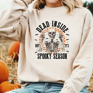 May include: A beige sweatshirt with a Halloween-themed graphic. The graphic features a skeleton with peace signs, surrounded by bats and the text "DEAD INSIDE BUT IT'S SPOOKY SEASON". Pumpkins are visible in the background.