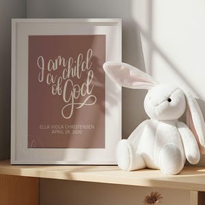 Può includere: Una stampa incorniciata con il testo "I am a child of God" in elegante scrittura, con il nome "Ella Viola Christensen" e la data "April 29, 2020" sotto. Un coniglietto di peluche bianco con orecchie rosa è accanto alla cornice.