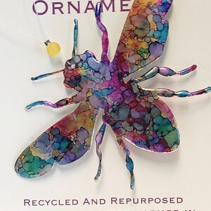 May include: A colourful bee-shaped ornament made from a recycled aluminium can. The ornament is painted with a variety of colours, including blue, green, purple, and pink. The text "RECYCLED AND REPURPOSED NO BEVERAGES WERE HARMED IN THE MAKING OF THIS ORNAMENT" is printed on the white background.