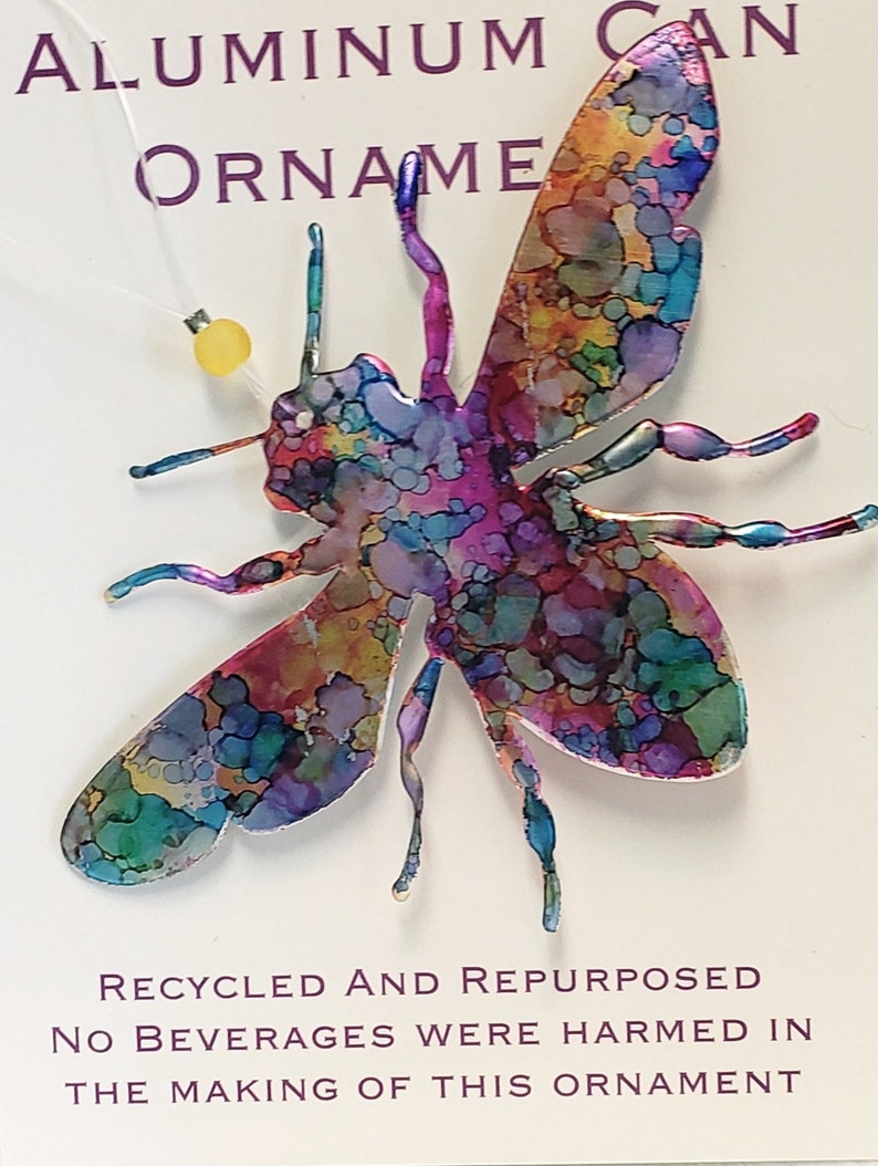 May include: A colourful bee-shaped ornament made from a recycled aluminium can. The ornament is painted with a variety of colours, including blue, green, purple, and pink. The text "RECYCLED AND REPURPOSED NO BEVERAGES WERE HARMED IN THE MAKING OF THIS ORNAMENT" is printed on the white background.