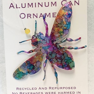 May include: A colourful bee ornament made from a recycled aluminium can. The bee is painted with a variety of colours, including blue, green, purple, and yellow. The ornament is hanging from a string with a small yellow bead. The text on the card reads "Aluminium Can Ornament, Recycled and Repurposed, No Beverages Were Harmed in the Making of This Ornament."