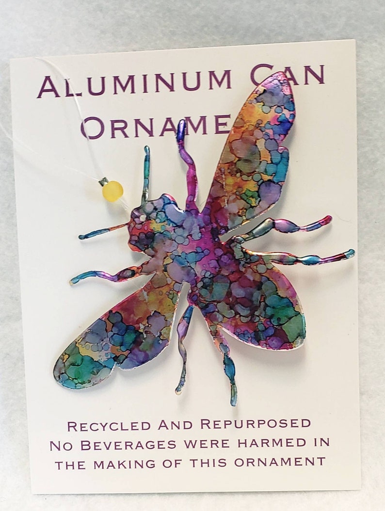 May include: A colourful bee ornament made from a recycled aluminium can. The bee is painted with a variety of colours, including blue, green, purple, and yellow. The ornament is hanging from a string with a small yellow bead. The text on the card reads "Aluminium Can Ornament, Recycled and Repurposed, No Beverages Were Harmed in the Making of This Ornament."