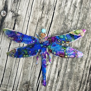May include: A blue, purple, and green dragonfly with a turquoise bead in the center. The dragonfly is made of metal and has a detailed, abstract design.
