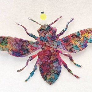 May include: A colourful, abstract bee ornament with a light yellow dot on top. The bee is made of metal and has a detailed, intricate design.