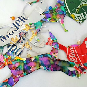 May include: A collection of colourful, abstract bird ornaments made from recycled aluminium cans. The birds are painted in shades of blue, green, purple and pink. The cans have logos for Budweiser, Michelob and a green can with a heart shape.