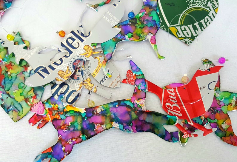 May include: A collection of colourful, abstract bird ornaments made from recycled aluminium cans. The birds are painted in shades of blue, green, purple and pink. The cans have logos for Budweiser, Michelob and a green can with a heart shape.