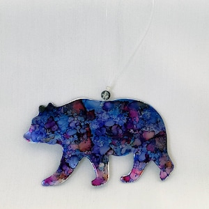 May include: A decorative bear ornament with a colorful abstract design in shades of blue, purple, and pink. The bear is walking, with a silver outline and a clear string for hanging. A small bead is attached to the string.
