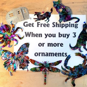 May include: A collection of colourful metal ornaments, including a butterfly, dragonfly, octopus, crab, dinosaur, cat, and a camper. The ornaments are hanging from strings and have a watercolour-like paint finish. The text "Get Free Shipping When you buy 3 or more ornaments" is written on a white background.