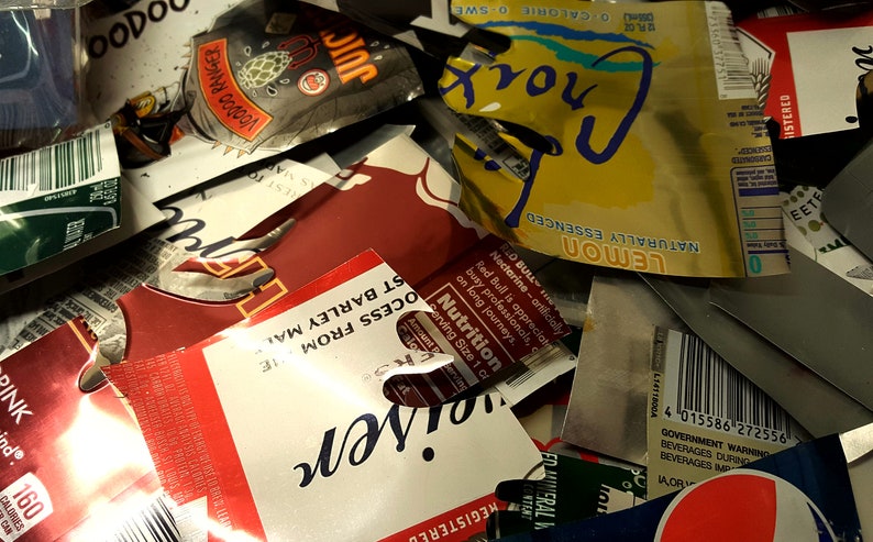 May include: A close-up of a pile of crushed aluminium cans, including a red can with the word "Red Bull" and a yellow can with the words "Naturally Essenced Lemon Croix".