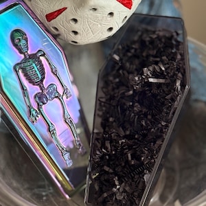 Op de afbeelding: Twee kistvormige containers. Eén iriserend met een skeletafbeelding, de andere gevuld met zwart gescheurd papier. Een wit masker met rode accenten staat op de achtergrond.