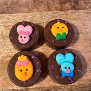 Puede incluir: Cuatro galletas cubiertas de chocolate decoradas con dise&ntilde;os tem&aacute;ticos de Pascua. Cada galleta presenta un dise&ntilde;o diferente: un conejito rosa, un pollito amarillo, un huevo amarillo y un conejito azul. Chispas moradas rodean cada galleta.