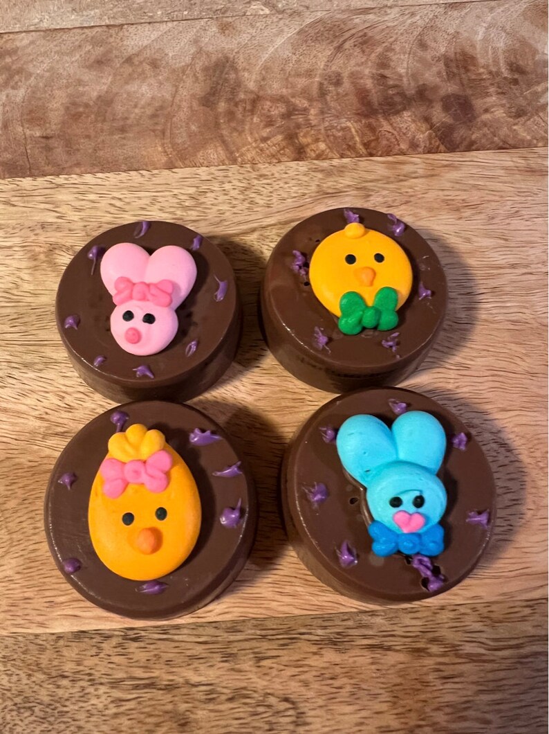 Puede incluir: Cuatro galletas cubiertas de chocolate decoradas con dise&ntilde;os tem&aacute;ticos de Pascua. Cada galleta presenta un dise&ntilde;o diferente: un conejito rosa, un pollito amarillo, un huevo amarillo y un conejito azul. Chispas moradas rodean cada galleta.