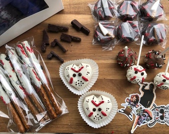 Könnte beinhalten: Eine Auswahl an Halloween-Leckereien, darunter in weiße Schokolade getauchte Brezelstäbchen, die mit rotem und weißem Zuckerguss verziert sind, herzförmige Schokoladen-Leckereien mit einem weißen Schokoladen-Masken-Design und schokoladenüberzogene Cake Pops mit roten und weißen Streuseln.