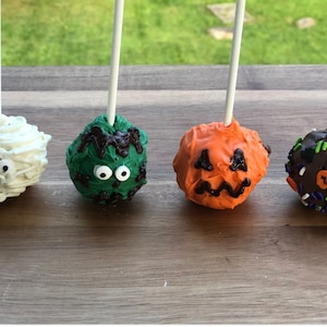 May include: Four Halloween-themed cake pops on sticks. The cake pops are decorated with white, green, orange, and brown frosting. The white cake pop is decorated to look like a mummy, the green cake pop is decorated to look like a monster, the orange cake pop is decorated to look like a pumpkin, and the brown cake pop is decorated with sprinkles.