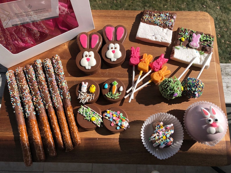 Puede incluir: Una tabla de madera exhibe una variedad de dulces de Pascua. Pretzels cubiertos de chocolate con chispas, chocolates con forma de conejito, malvaviscos con chocolate y cake pops. Tambi&eacute;n hay piruletas con forma de conejito y un cake pop rosa con una cara de conejito.