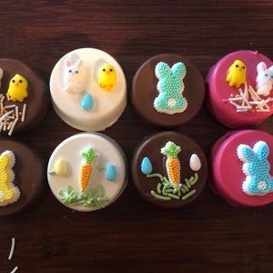 Puede incluir: Ocho galletas s&aacute;ndwich cubiertas de chocolate decoradas con dise&ntilde;os de Pascua. Las galletas presentan figuras de conejitos y pollitos, zanahorias y huevos, con chispas de colores. Las galletas est&aacute;n sobre una superficie de madera oscura.