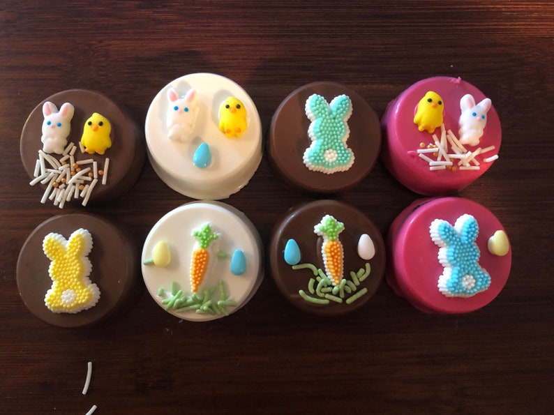 Puede incluir: Ocho galletas s&aacute;ndwich cubiertas de chocolate decoradas con dise&ntilde;os de Pascua. Las galletas presentan figuras de conejitos y pollitos, zanahorias y huevos, con chispas de colores. Las galletas est&aacute;n sobre una superficie de madera oscura.