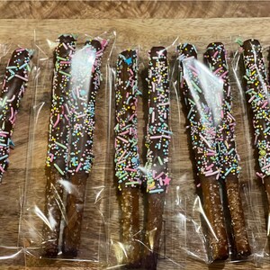 Puede incluir: Varias barras de pretzel cubiertas de chocolate, decoradas con chispas de colores, est&aacute;n empaquetadas individualmente en pl&aacute;stico transparente. Los pretzels est&aacute;n dispuestos sobre una superficie de madera, mostrando un dulce.
