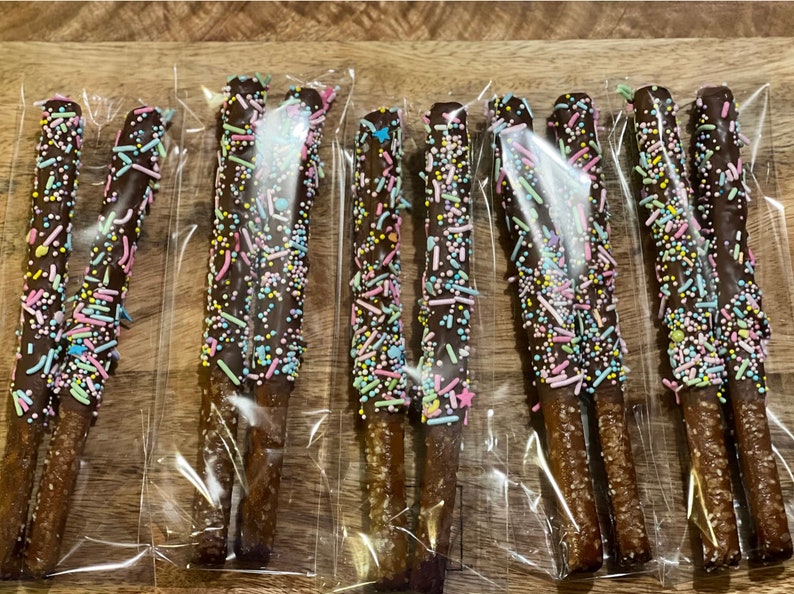 Puede incluir: Varias barras de pretzel cubiertas de chocolate, decoradas con chispas de colores, est&aacute;n empaquetadas individualmente en pl&aacute;stico transparente. Los pretzels est&aacute;n dispuestos sobre una superficie de madera, mostrando un dulce.