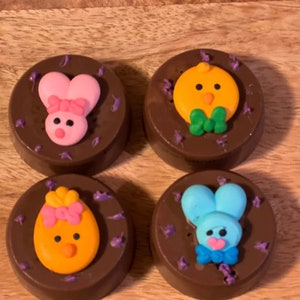 Puede incluir: Cuatro golosinas cubiertas de chocolate con decoraciones tem&aacute;ticas de Pascua. Dos presentan dise&ntilde;os de conejitos rosas, uno con un lazo. Los otros dos tienen dise&ntilde;os de pollitos amarillos, uno con un lazo. Todos est&aacute;n decorados con detalles morados.