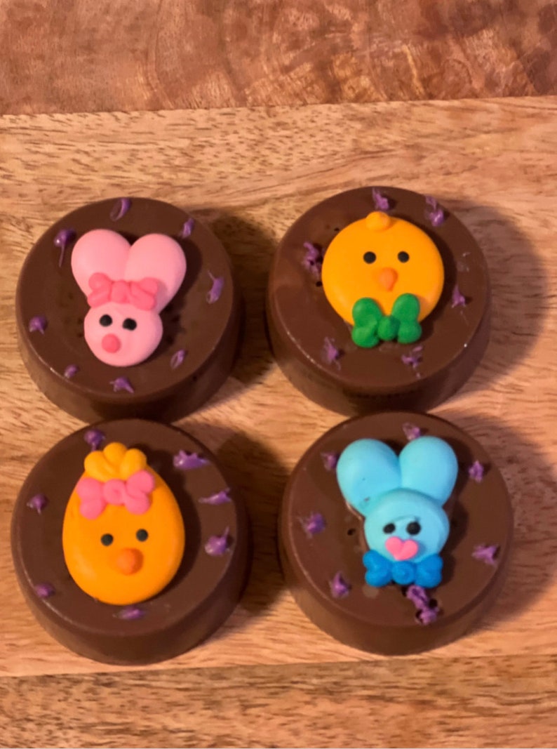 Puede incluir: Cuatro golosinas cubiertas de chocolate con decoraciones tem&aacute;ticas de Pascua. Dos presentan dise&ntilde;os de conejitos rosas, uno con un lazo. Los otros dos tienen dise&ntilde;os de pollitos amarillos, uno con un lazo. Todos est&aacute;n decorados con detalles morados.