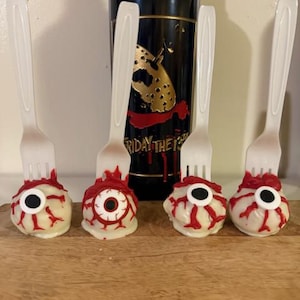 May include: Four white chocolate eyeball cake pops with red icing and black pupils on wooden surface with a bottle of wine in the background.