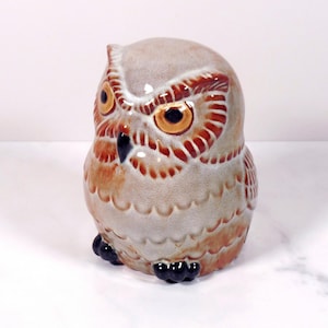 May include: A ceramic owl figurine featuring a glossy finish with a color palette of light gray, tan, and brown. The owl has prominent orange eyes with black pupils, a dark beak, and black feet, with detailed feather patterns etched into the surface.