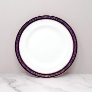 May include: White ceramic plate with a navy blue and gold leaf border. The plate has a wide navy blue band with a gold leaf pattern and a gold dotted inner border. The center of the plate is white.