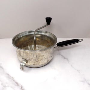 May include: A vintage metal food mill featuring a round basin with a weathered silver finish, a black plastic handle on the side, and a central rotating crank mechanism topped with a black knob for manual operation of the internal food processing blade.