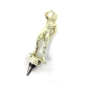 May include: Gold-colored bottle stopper shaped like a person standing on a platform. The person is holding their head with one hand and has a sculpted, muscular physique.