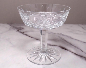 May include: A clear crystal champagne coupe glass. The bowl has a diamond-cut pattern, and the stem is a solid, clear column. The base is a circular design with radiating lines. The glass is designed for serving champagne or other sparkling wines.