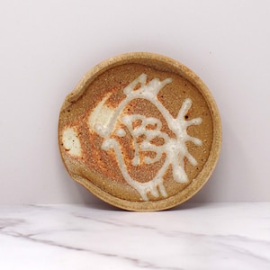 May include: A small, round, ceramic spoon rest with a white design of a stylized bird or flower. The rest is a light brown color with a speckled glaze.