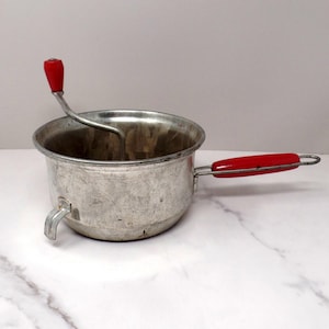 May include: A vintage metal food mill featuring a round basin with a textured surface, a side handle with a red grip, and a top-mounted rotating crank handle with a red knob. The device includes a small metal support leg on the exterior base.