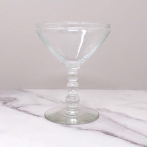May include: A clear glass martini glass with a beaded stem. The glass is sitting on a white marble surface.