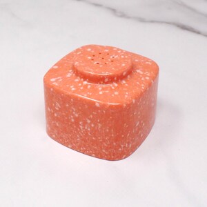 May include: A coral-colored, square-shaped salt shaker with a speckled pattern. The top features a smaller, circular section with several holes for dispensing salt. The shaker is made of a solid material and has a smooth, polished finish.