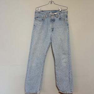 May include: Light wash denim jeans hanging on a metal hanger. The jeans have a classic five-pocket design, a button fly, and a straight leg fit. The denim has a faded, worn appearance.