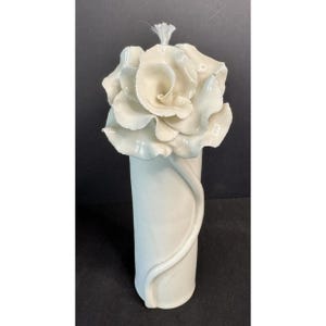 May include: A white ceramic vase with a large rose bloom at the top. The vase has a cylindrical shape with a decorative, curved accent. The rose is detailed with multiple petals, and the overall design is elegant.