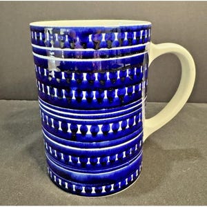 May include: A cylindrical ceramic mug featuring a vibrant cobalt blue and white pattern. The design consists of horizontal bands filled with repeating teardrop or petal-like shapes, separated by thin white lines, and includes a plain white handle.