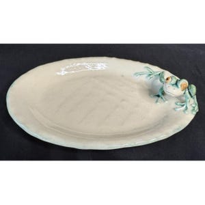 May include: Off-white ceramic serving platter with a frog accent. The oval-shaped dish has a light blue rim and a frog figurine on one end, featuring green and brown details. The frog is perched on the edge of the platter.